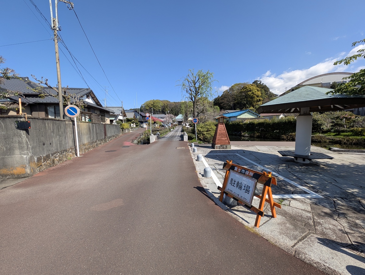 泉の郷湧水公園の駐輪場と、屋根付きの休憩スペース（東屋）。奥には医王寺へ続く道が見える風景。